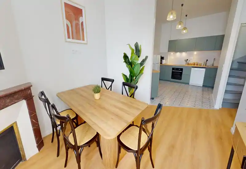 Zona pranzo luminosa e arredata con un tavolo in legno e quattro sedie vicino a una cucina a vista; toni naturali, una pianta decorativa e un quadro creano un'atmosfera accogliente.