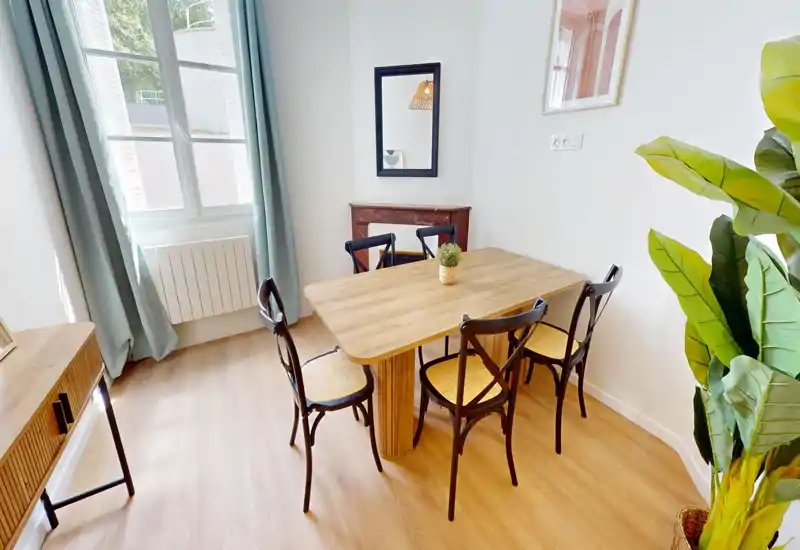 Zona pranzo luminosa e curata con tavolo in legno e quattro sedie, luce naturale da una grande finestra e pianta decorativa che dà freschezza.