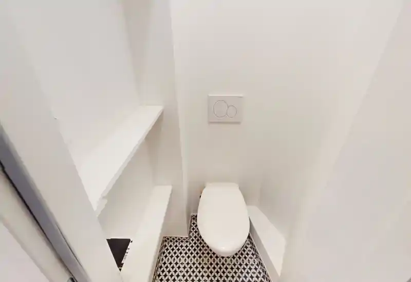 Bagno/WC compatto e moderno con piastra di scarico a muro e pavimento con piastrelle decorative. Pareti bianche e mensole integrate donano un aspetto pulito e minimale.