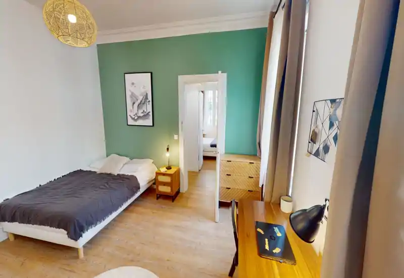 Chambre lumineuse et bien aménagée avec lit double, table de chevet et bureau près de la fenêtre. Textiles doux, lampe de chevet et mur vert apportent une ambiance calme et accueillante, parfaite pour des photos de location.