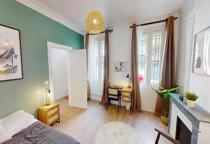 Chambre lumineuse et moderne avec un lit confortable, une table de chevet et un petit bureau près de grandes fenêtres laissant entrer la lumière naturelle. Mur vert d'accent et suspension en rotin pour une atmosphère chaleureuse.