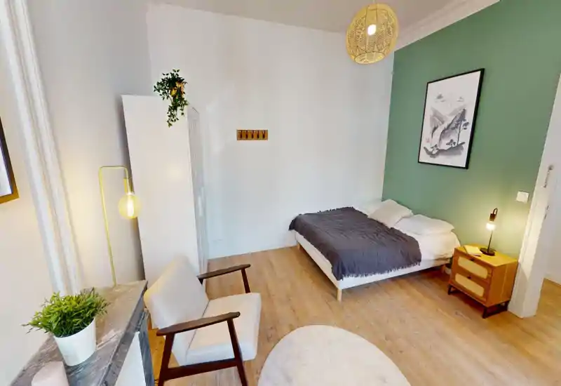 Chambre lumineuse et aménagée avec lit double devant un mur vert, table de chevet avec lampe, armoire et fauteuil confortable — décor cosy et moderne, parfait pour une annonce locative.
