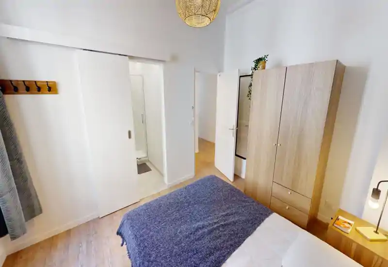 Chambre lumineuse et bien mise en scène avec un grand lit couvert d’un plaid bleu, une armoire et une table de chevet en bois avec lampe — style moderne et chaleureux.