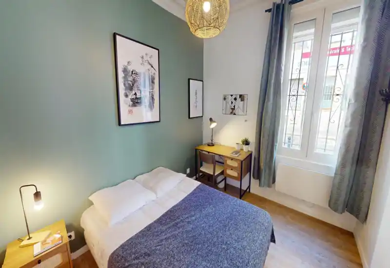 Chambre compacte et élégante avec un lit double, une lampe de chevet et un bureau en bois près d'une fenêtre lumineuse — mur vert doux et tableaux encadrés pour une ambiance chaleureuse.