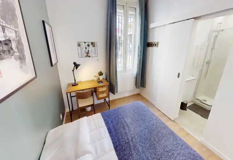 Chambre compacte et lumineuse avec lit visible, bureau en bois et fenêtre apportant de la lumière naturelle ; décoration soignée avec un chemin de lit bleu et une lampe de bureau.