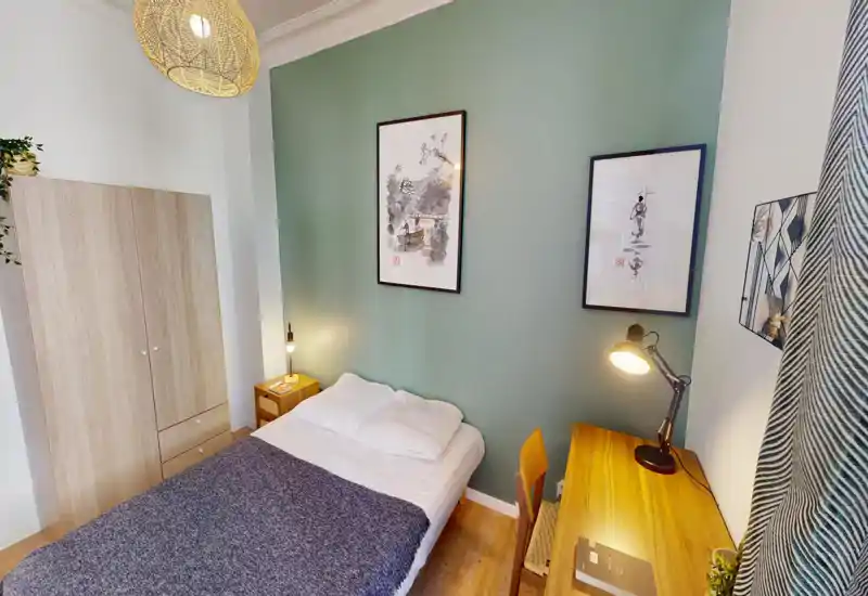 Chambre lumineuse et décorée avec lit double, tables de chevet et bureau en bois. Mur vert d'accent, tableaux encadrés et lampes chaleureuses offrent une ambiance cosy et moderne, parfaite pour une location courte durée.