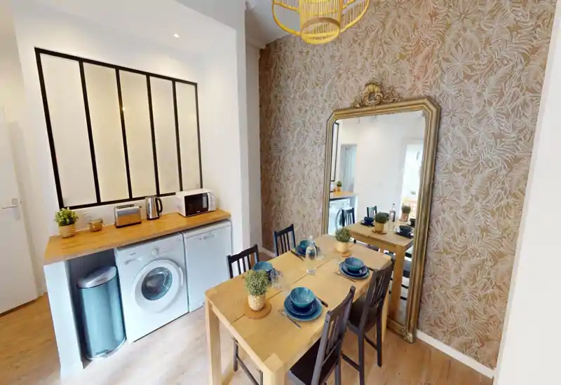 Accogliente zona pranzo ben arredata con tavolo in legno per quattro persone, piante decorative e carta da parati calda; un grande specchio ornamentale amplia visivamente lo spazio.
