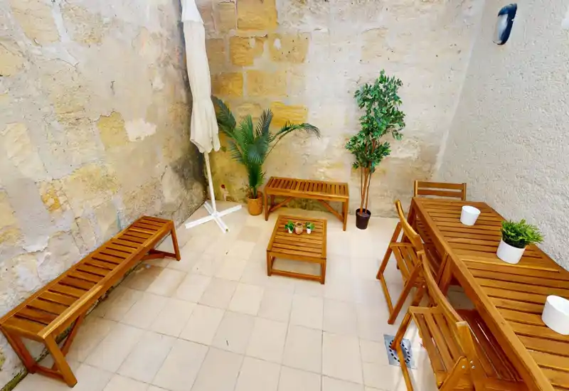 Cortile soleggiato con mobili da esterno in legno — panche, tavolo con sedie e piante in vaso creano una terrazza privata accogliente, ideale per caffè mattutini o piccoli incontri.