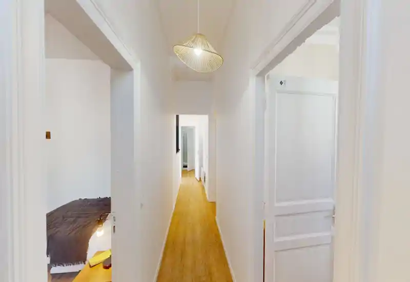 Corridoio luminoso e stretto con pavimento in legno, pareti bianche e una lampada a sospensione; porte aperte verso le stanze adiacenti e una camera parzialmente visibile a sinistra.