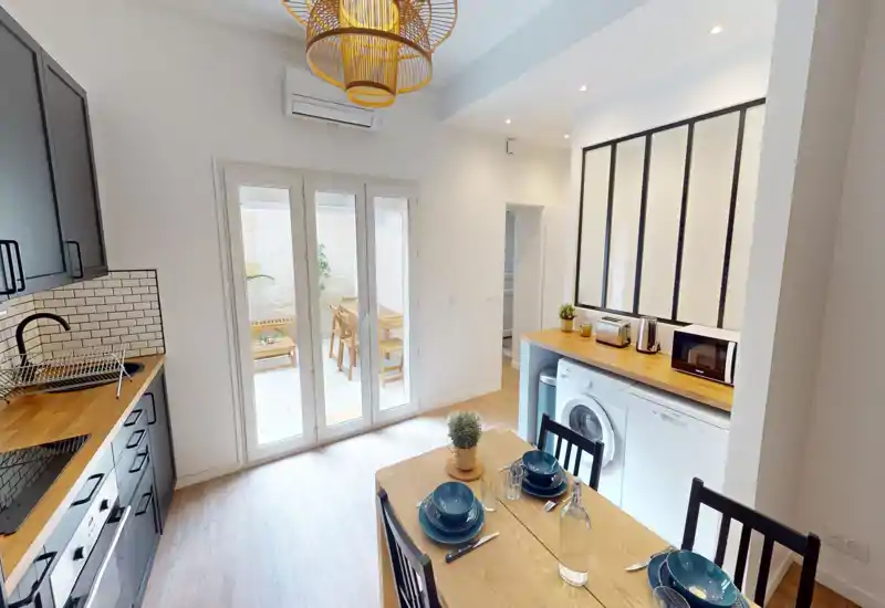 Cucina open-space luminosa con piani in legno e un piccolo tavolo da pranzo per quattro persone; porte in vetro si affacciano su un patio soleggiato. Elettrodomestici moderni, rivestimento a mattoncini e lampade a sospensione donano un ambiente accogliente e funzionale, ideale per affitti brevi.