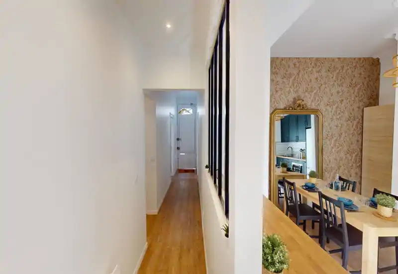 Zona pranzo luminosa e arredata con tavolo in legno apparecchiato, carta da parati decorativa e uno specchio grande che riflette la cucina adiacente — perfetta per pasti conviviali.