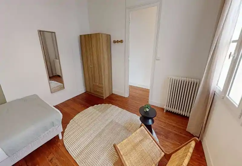 Camera da letto luminosa e accogliente con pavimenti in legno, un letto singolo, un armadio e una sedia in vimini — ideale per un soggiorno confortevole con luce naturale.