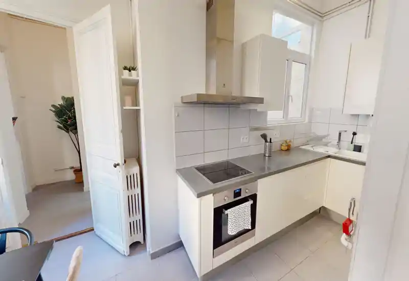 Cucina moderna compatta con forno integrato e piano a induzione, pensili chiari e finestra che illumina l'ambiente. Piano di lavoro pulito e piastrelle neutre per un'area cottura luminosa e funzionale.