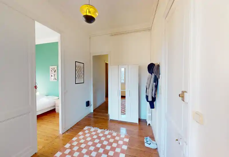 Ingresso luminoso con pavimento in legno, tappeto a quadretti e un armadio; la porta aperta conduce a una camera con parete verde.