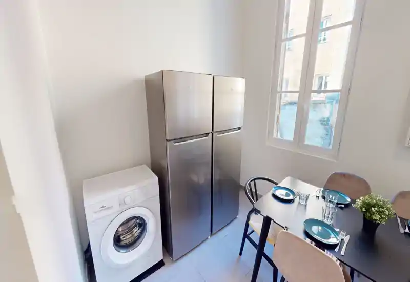 Cucina compatta e moderna con frigorifero alto in acciaio, lavatrice e un piccolo tavolo da pranzo per quattro. La luce naturale dalla grande finestra illumina lo spazio pulito e contemporaneo.