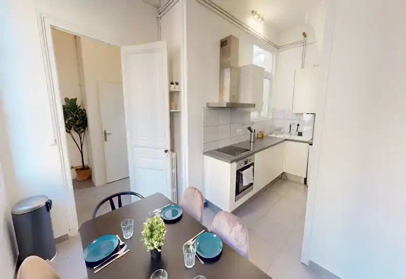 Cucina open-space compatta e luminosa con piano cottura moderno, forno e bancone a L; un tavolo da pranzo apparecchiato per quattro in primo piano, ideale per un annuncio che valorizzi uno spazio funzionale e curato.
