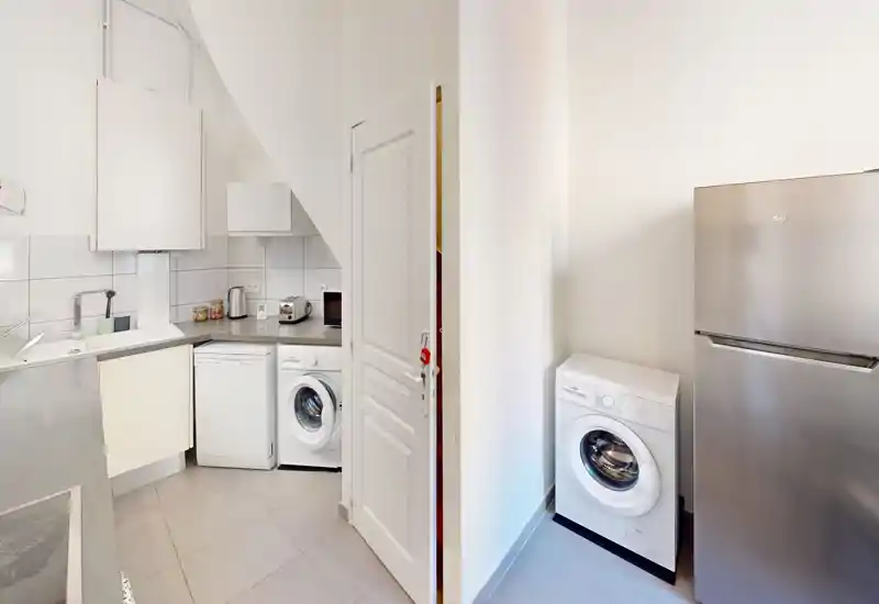 Cucina moderna compatta con elettrodomestici integrati tra cui lavastoviglie, lavatrice e frigorifero in acciaio inox, piastrelle chiare e disposizione a L efficiente.