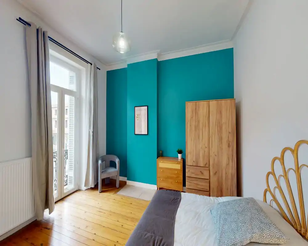 Chambre lumineuse et bien mise en scène avec un mur d'accent bleu canard, parquet en bois et lit double partiellement visible. La lumière naturelle entre par les portes-fenêtres et le mobilier en bois apporte une atmosphère chaleureuse.