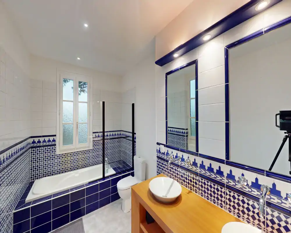 Salle de bains lumineuse carrelée avec baignoire, WC et meuble-lavabo en bois surmonté d'une vasque et d'un grand miroir.