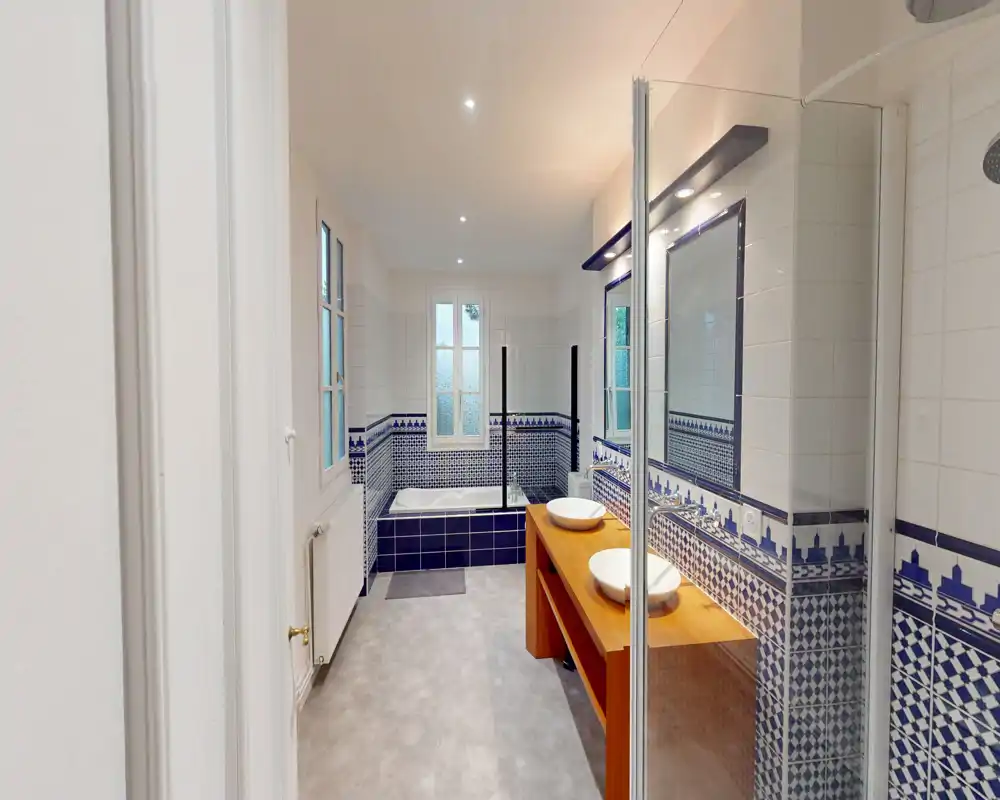 Salle de bains lumineuse et spacieuse avec une baignoire carrelée, deux vasques sur un meuble en bois et une paroi de douche en verre; carrelage bleu décoratif.