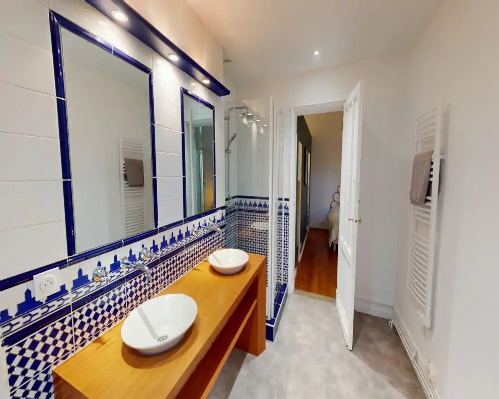 Salle de bains lumineuse et soignée avec deux vasques sur meuble en bois, carreaux décoratifs bleu et blanc et douche vitrée.