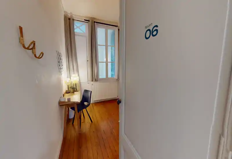 Petite pièce de type chambre avec parquet, un bureau étroit et une chaise près d'une grande fenêtre ; lampes chaudes et rideaux neutres pour un coin accueillant.