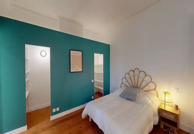 Chambre lumineuse et stylée avec une tête de lit en rotin décorative, literie blanche et lampe de chevet. Le mur d'accent turquoise encadre deux alcôves avec étagères et le parquet apporte une ambiance chaleureuse.