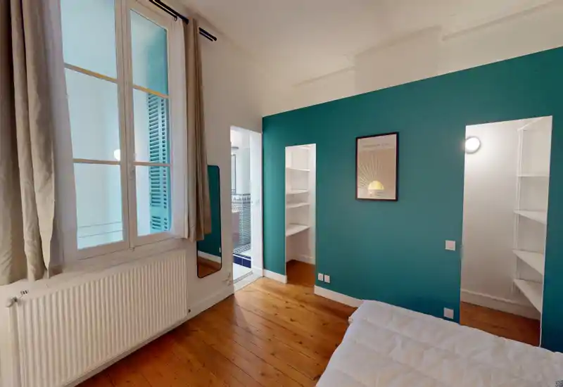 Coin chambre lumineux et bien agencé avec un mur d'accent bleu-vert, parquet et lit partiellement visible. Étagères intégrées et accès à une salle de bains ou dressing.