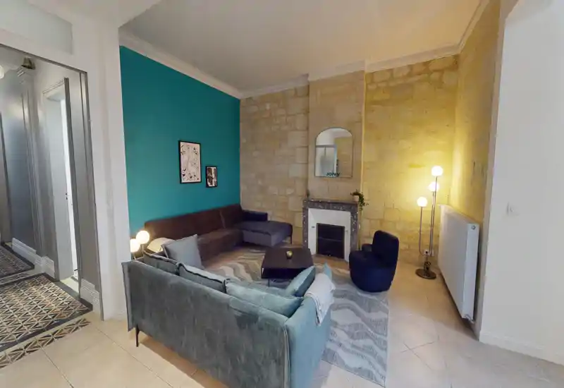 Soggiorno luminoso e di design con comodo divano angolare, poltrona, tappeto decorato e camino decorativo su parete in pietra a vista.