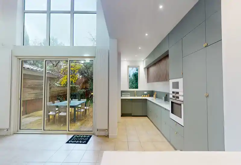 Cucina moderna open space con pensili verde salvia, elettrodomestici integrati e ampia porta scorrevole che si apre su un patio esterno.