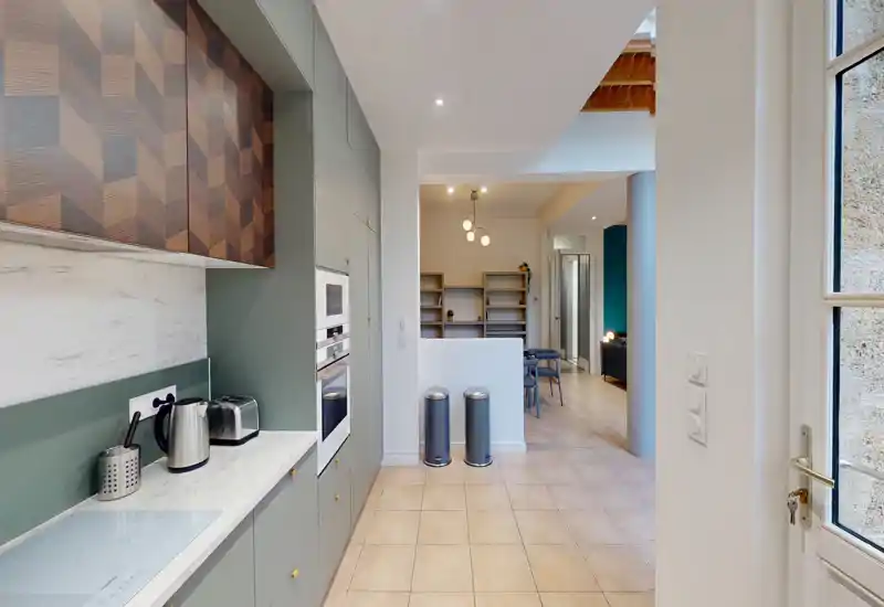 Cucina moderna e compatta con elettrodomestici integrati, piano di lavoro effetto marmo e pensili verde salvia; la luce naturale dalla porta vetrata e gli accenti in legno rendono l'ambiente caldo e funzionale.