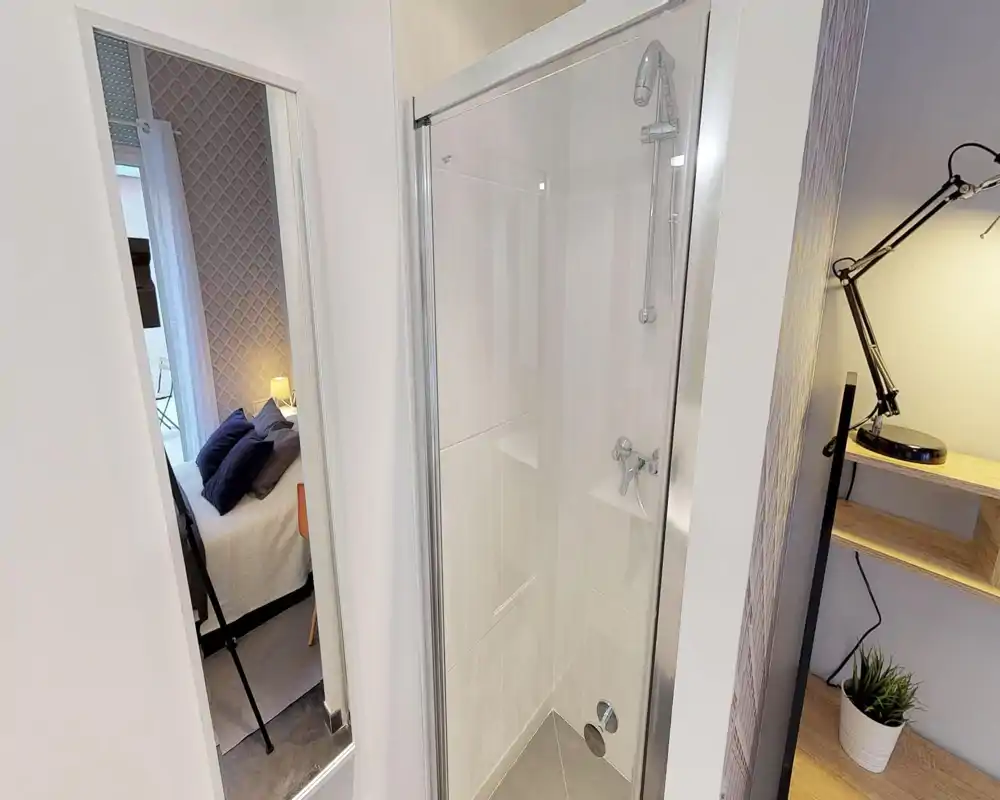 Cosy bedroom corner visible through a doorway: a neatly made bed with cushions, bedside lamp and patterned headboard create a welcoming sleeping area partially seen from the corridor.