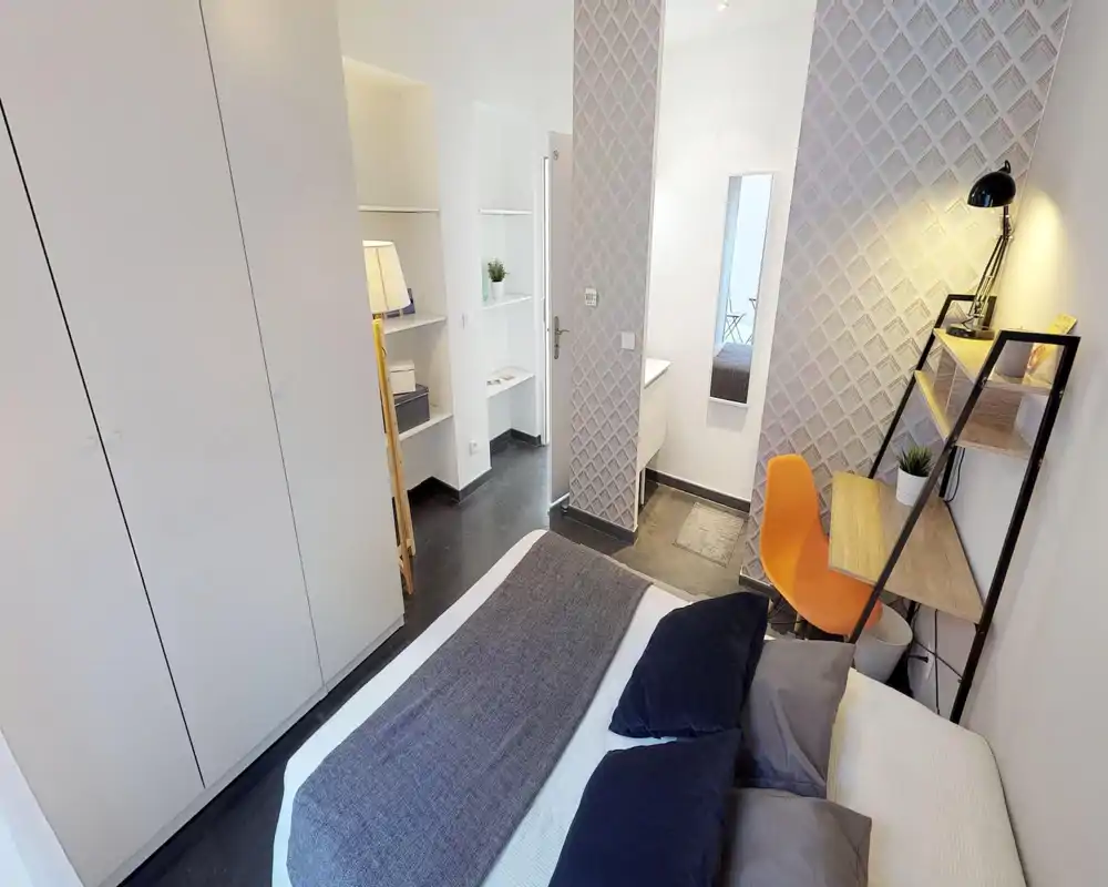 Bright, modern bedroom with a large bed in the foreground, built-in wardrobe, and a small desk with shelving. Neutral tones and tasteful textiles create a calm, welcoming space ideal for short stays.
