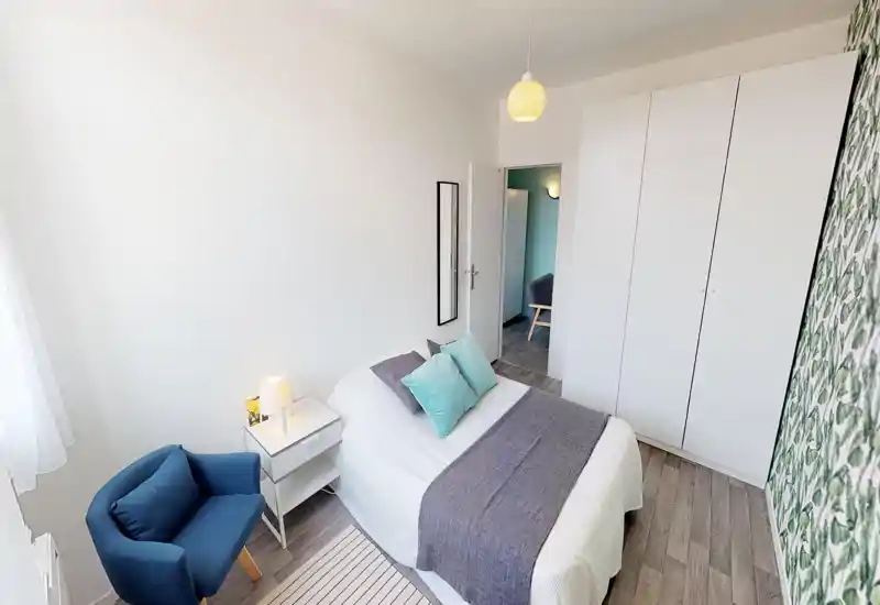 Camera da letto moderna e luminosa con letto matrimoniale ben rifatto, comodino con lampada, poltroncina blu e ampia armadiatura bianca. Cuscini turchesi e plaid grigio aggiungono colore; finestra grande assicura luce naturale.