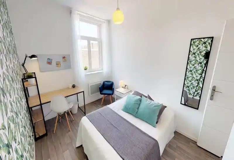 Camera da letto luminosa e moderna con letto ben rifatto, cuscini color turchese e una scrivania in legno vicino alla finestra; carta da parati botanica e specchio a figura intera aggiungono stile.