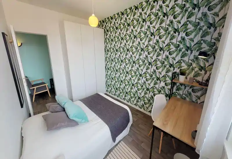 Camera da letto luminosa e di stile con un comodo letto matrimoniale, carta da parati botanica e una piccola scrivania vicino alla finestra. Finiture moderne e cuscini coordinati rendono l’ambiente accogliente e ben allestito.