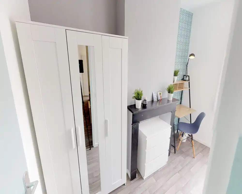 Coin de chambre lumineux avec une grande armoire blanche, une petite commode et un bureau soigné avec chaise — minimaliste, bien éclairé et décoré de petites plantes.