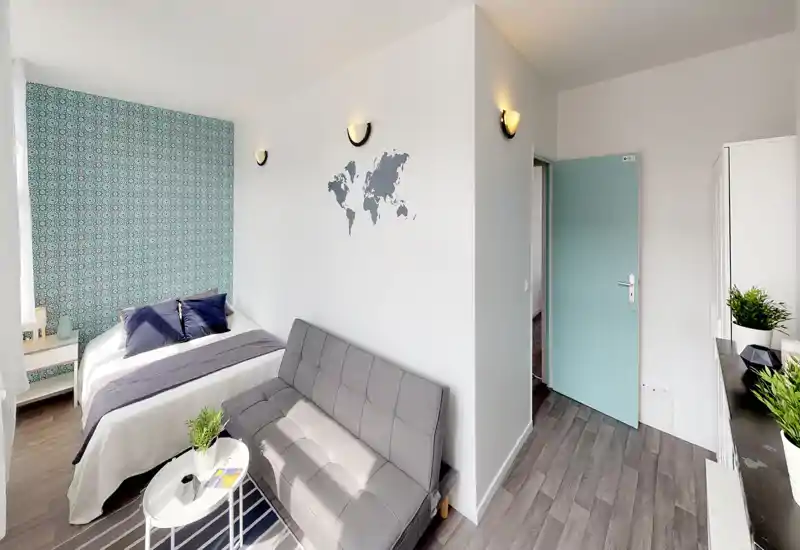 Chambre-studio claire et tendance avec un lit double bien fait, un mur à motif et un petit canapé gris — idéale pour courts séjours. Lumière naturelle, décoration soignée et agencement fonctionnel rendent l’espace chaleureux.