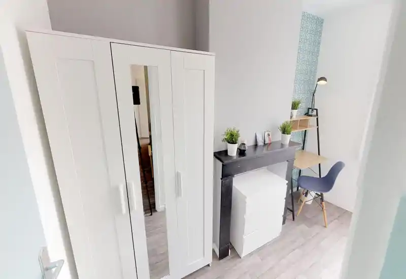 Coin de chambre lumineux avec une grande armoire blanche, une petite commode et un bureau soigné avec chaise — minimaliste, bien éclairé et décoré de petites plantes.