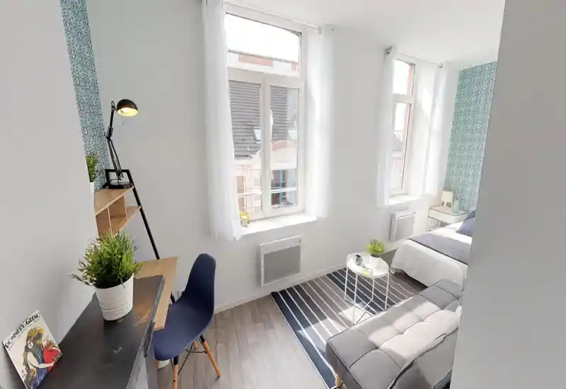 Chambre lumineuse et bien aménagée avec lit double, banquette et petit espace bureau près de la fenêtre. Murs blancs, papier peint à motifs et plantes apportent une atmosphère fraîche et accueillante.