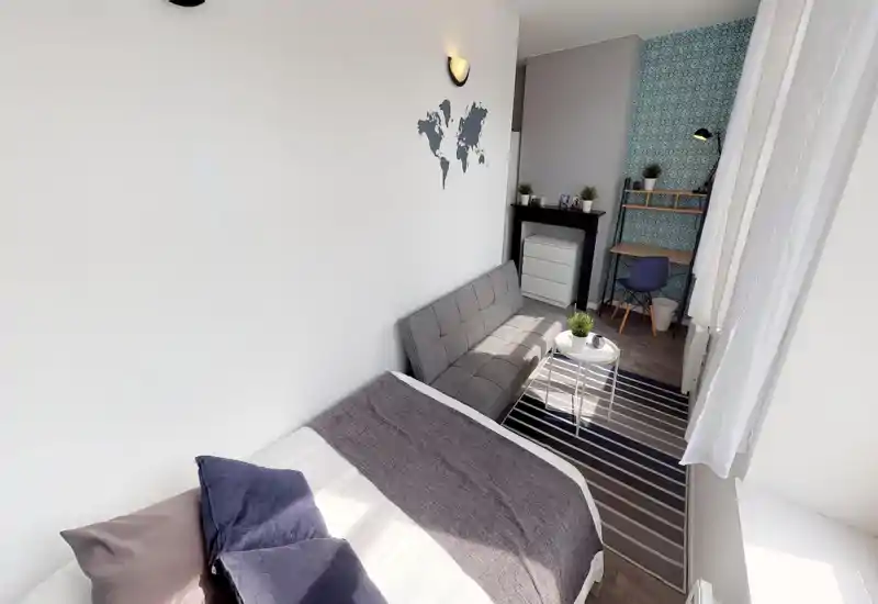 Chambre lumineuse et bien décorée avec grand lit visible, petit canapé, table basse et coin bureau devant un mur à motif — idéale pour une personne ou un couple.