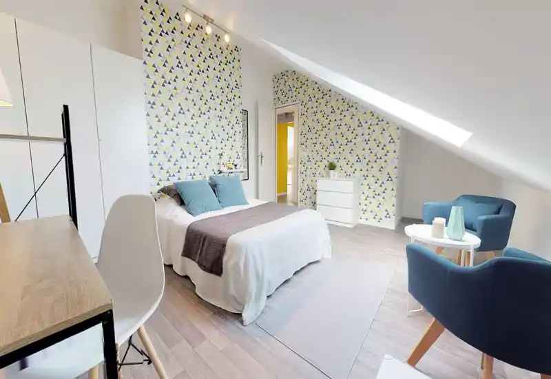 Bright, stylish attic bedroom with patterned feature wall, a neatly made double bed with turquoise cushions, a cozy seating area and skylight providing ample natural light.