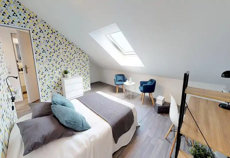 Bright attic bedroom with a comfortable double bed, patterned feature wall and a skylight; sitting area with two blue armchairs and a small table adds cozy appeal.