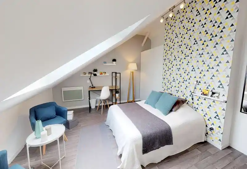 Bright, styled attic bedroom with a double bed, patterned accent wall and a cozy reading corner with armchair and desk — ideal for a comfortable stay.