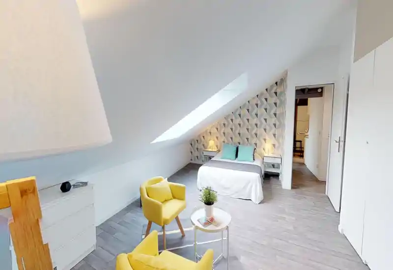 Chambre mansardée lumineuse avec un lit double bien fait, un mur d'accent géométrique et deux tables de chevet avec lampes. Coin salon avec deux fauteuils jaunes et une petite table, idéal pour se détendre.