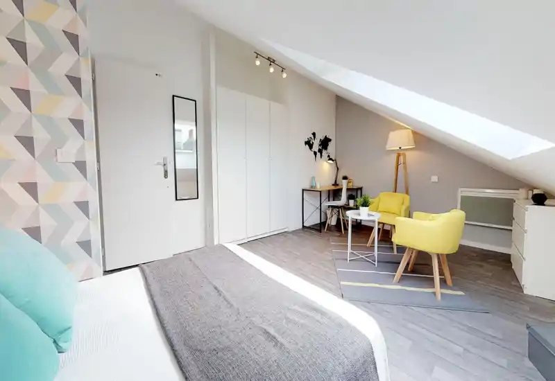 Chambre mansardée lumineuse et moderne avec lit au premier plan, coin salon avec fauteuils jaunes, bureau et fenêtre de toit apportant une grande luminosité.