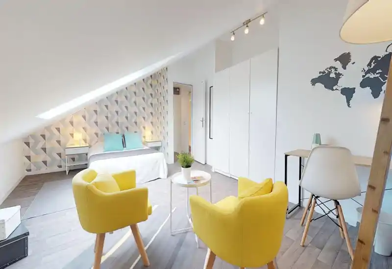 Chambre mansardée lumineuse et moderne avec lit double sous une fenêtre de toit, mur d'accent géométrique pastel, tables de chevet avec lampes et coin salon confortable avec deux fauteuils jaunes.