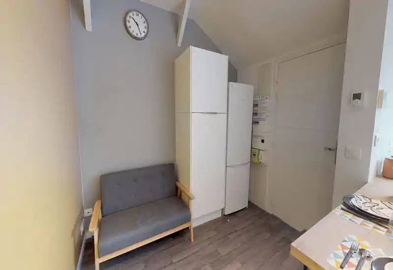 Petit espace salon avec un canapé deux places, accents en bois clair et un réfrigérateur contre le mur ; lumineux, soigné et adapté à un studio.