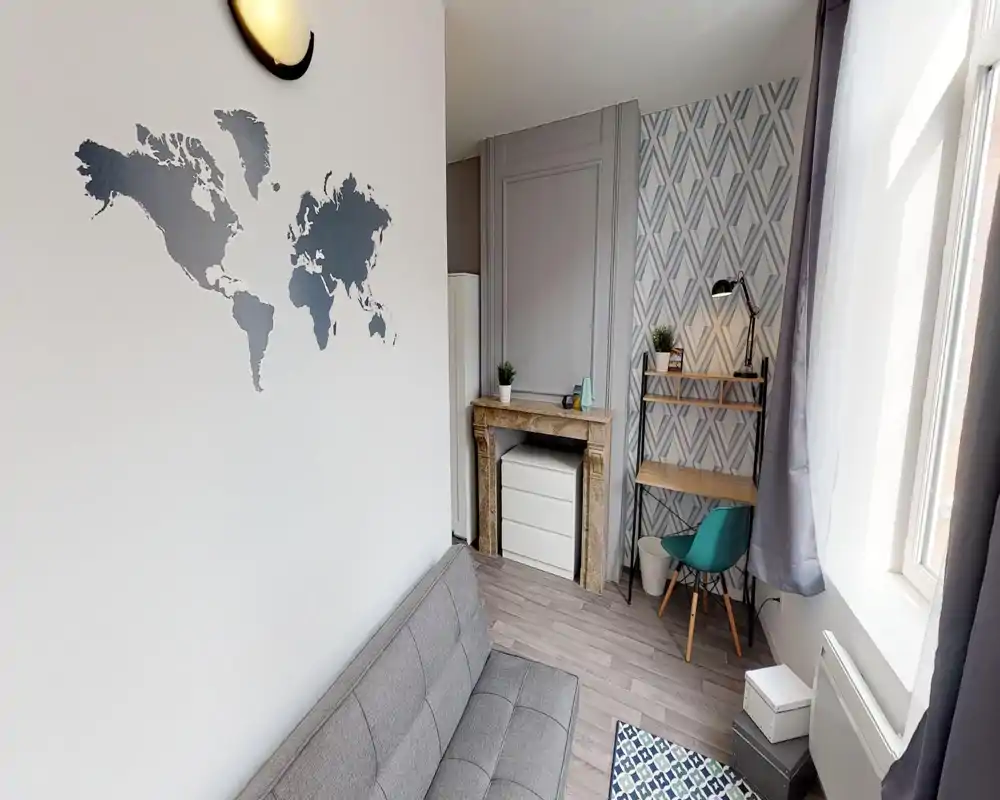 Petit salon moderne et lumineux avec canapé gris, tapis à motifs et bureau compact près de la fenêtre ; sticker mural et plantes apportent du caractère.