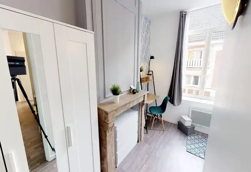 Petit coin chambre bien éclairé avec bureau et armoire. La pièce est soignée, décorée d’une cheminée décorative, d’une plante et d’une chaise près de la fenêtre, parfait pour un espace de travail lumineux.